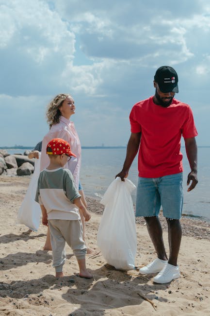 A man with dark skin, wearing a black baseball cap, red t-shirt, light blue distressed denim shorts, and white sneakers, is standing on a sandy beach holding a large white plastic rubbish bag in his right hand. Next to him, a young boy wearing a red cap with a yellow pattern, a light-colored t-shirt, and beige shorts is also holding a white bag, possibly for collecting litter. A woman with curly blonde hair, dressed in a light pink top, stands behind them, observing the scene. In the background, the shoreline extends to the horizon with calm water, some rocks, and an overcast sky filled with various shades of clouds. The scene appears to depict a community or family beach clean-up, emphasizing activities related to waste collection and environmental care, similar to services provided by Rubbish Clearance Docklands, supporting independent waste disposal outside of local authorities.