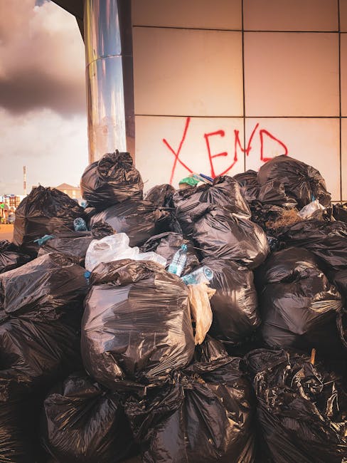 An outdoor scene showing a pile of mixed rubbish on a gravel surface, including several black plastic trash bags, a yellow plastic storage container, a worn car tyre, and various smaller debris. The rubbish is situated against a low stone wall made of irregularly shaped, rough-textured stones, with some greenery, including bushes and small trees, visible behind the wall. In the background, there is a fence and a large structure with a curved roof, possibly a sports or industrial facility, along with utility poles and overhead power lines against a partly cloudy sky with patches of blue. The scene captures a collection of discarded waste likely awaiting removal, demonstrating an environment that could benefit from private waste management services such as those offered by Rubbish Clearance Docklands, highlighting the importance of proper rubbish disposal and the role of independent collection options.