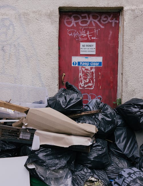 A stack of black trash bags filled with waste material piled in front of a red metal door set into a light-colored, textured concrete wall. The black bags appear to contain household rubbish and are stacked on top of each other, some resting on discarded flattened cardboard and packaging materials. The door features graffiti markings in white and pink spray paint, along with a small sign reading 'No Money' and a larger blue and white sign that says 'Keep Clear.' The scene suggests a privately managed waste collection area, with the waste awaiting removal, consistent with an independent rubbish disposal service such as those offered by Rubbish Clearance Docklands operating in the local area. The environment appears to be an alleyway or service access point, with no visible surrounding greenery or outdoor features, and the overall atmosphere emphasizes the importance of proper rubbish handling and disposal alternatives outside local authority collection.