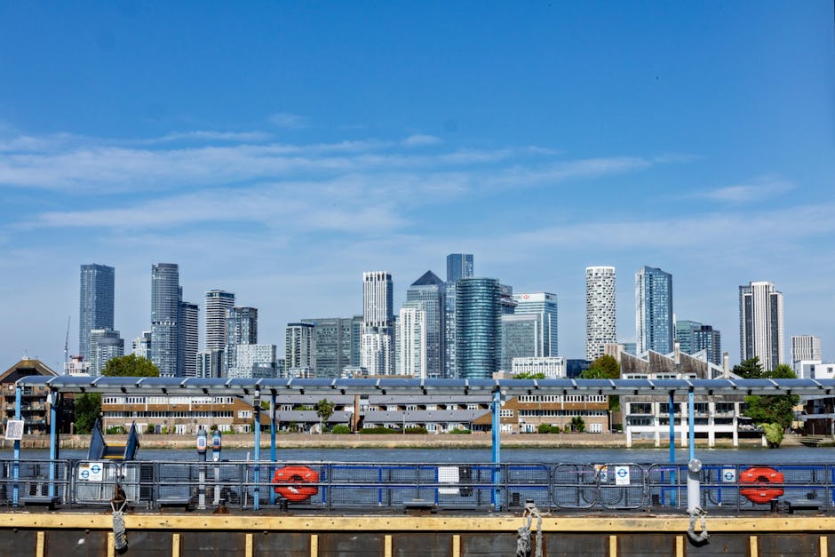 A view of a modern city skyline featuring numerous high-rise office buildings with glass facades reflecting sunlight, set against a clear blue sky with some wispy clouds. In the foreground, there is a riverside scene with a black and yellow mooring platform or dock area equipped with safety rails and various mooring fixtures, including red lifebuoys attached to the railings. Alongside the dock, there are small structures, possibly storage or maintenance sheds, with grey or metallic finishes. The scene is framed on the water's edge, and the middle ground shows a low-rise residential or commercial building with a flat roof, contrasting with the taller skyscrapers behind it. The environment suggests an urban riverside setting that might be relevant for private or alternative rubbish removal services, such as on-site clearance or waste handling away from traditional street collection, with the overall scene emphasizing a professional, neutral external perspective on a cityscape.