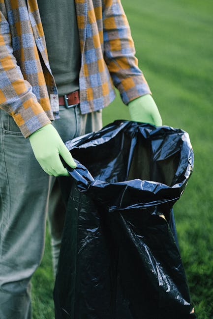A person standing outdoors on a grassy area, wearing a plaid shirt with yellow, blue, and gray tones over a green t-shirt, and light-colored trousers secured with a brown belt. They are holding open a large, black rubbish bag lined with a shiny, metallic inner layer. The individual is wearing bright green gloves for handling waste and is in the process of preparing the bag for rubbish collection, with their hands gripping the top edges. The scene suggests a waste management activity, possibly part of a community cleanup or private waste disposal effort, aligning with independent rubbish removal services. The background features an evenly lit, green, grassy field, providing a natural setting for environmentally conscious rubbish clearance. The image captures a professional and pragmatic approach to rubbish collection, emphasizing care in handling waste materials in an outdoor environment.