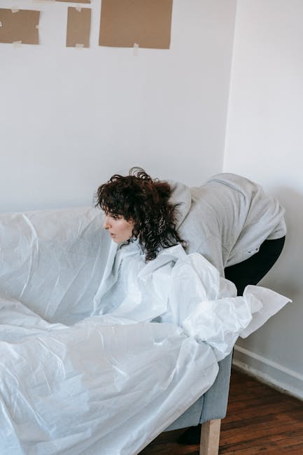 A woman with curly dark hair, dressed in a light gray hoodie and black trousers, is leaning forward over a white fabric covering a grey sofa, appearing to be arranging or removing the fabric. The scene is indoors, with a plain white wall in the background partially decorated with unpainted or patchy areas on the upper section. The grey sofa has a modern design with a smooth textured surface and wooden legs visible at the bottom right. The floor appears to be dark wood, contrasting with the white and neutral tones of the room. Natural light illuminates the space, creating a clear and neutral atmosphere. This image, while primarily depicting an interior activity, subtly relates to home clearance or organizing activities that might involve arranging or discarding household textiles, connecting to the broader context of rubbish removal or private waste handling services offered by Rubbish Clearance Docklands. The scene focuses on the careful handling of fabric in a domestic environment, indicative of a typical home clearance or organizing task.