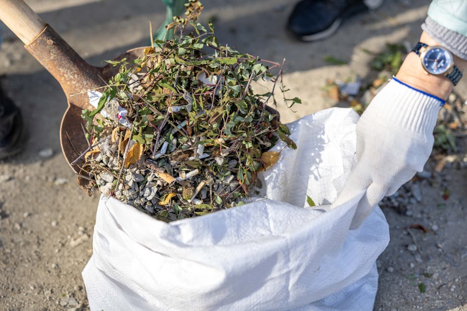 A person standing outdoors on a grassy area, wearing a plaid shirt with yellow, blue, and gray tones over a green t-shirt, and light-colored trousers secured with a brown belt. They are holding open a large, black rubbish bag lined with a shiny, metallic inner layer. The individual is wearing bright green gloves for handling waste and is in the process of preparing the bag for rubbish collection, with their hands gripping the top edges. The scene suggests a waste management activity, possibly part of a community cleanup or private waste disposal effort, aligning with independent rubbish removal services. The background features an evenly lit, green, grassy field, providing a natural setting for environmentally conscious rubbish clearance. The image captures a professional and pragmatic approach to rubbish collection, emphasizing care in handling waste materials in an outdoor environment.