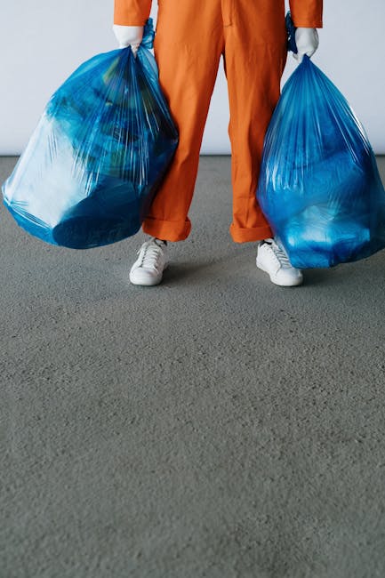 A person standing indoors on a grey carpeted floor, visible only from the waist down, wearing orange trousers and white sneakers, holding two large blue plastic rubbish bags filled with waste, one in each hand, with arms slightly bent. The bags are transparent in parts, showing crumpled recyclable materials inside. The background features a plain white wall, suggesting an indoor setting for waste collection or clearance. The scene exemplifies private rubbish disposal and on-site clearance activities, with the individual ready for waste removal services such as those offered by Rubbish Clearance Docklands, providing an informal but clear view of waste collection and disposal procedures fitting within the context of independent rubbish removal efforts.