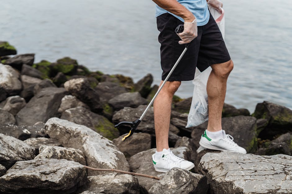 A person standing on a rocky shoreline near a body of water, wearing black shorts, a light blue T-shirt, and white sneakers with green accents. They are holding a litter picker in one hand and a partially filled transparent plastic bag in the other, suggesting they are engaged in a clean-up activity. The surrounding environment includes dark, rough-textured rocks with some patches of green moss or algae, and the water in the background appears calm with a soft, diffused light indicating an overcast day. The scene emphasizes outdoor environmental responsibility and the importance of maintaining natural coastal areas, aligning with private waste removal efforts often undertaken independently by individuals or small groups to clear litter and debris from shoreline environments. The individual’s stance and tools reflect active participation in rubbish collection, supporting the broader context of alternative waste handling beyond municipal collection services.