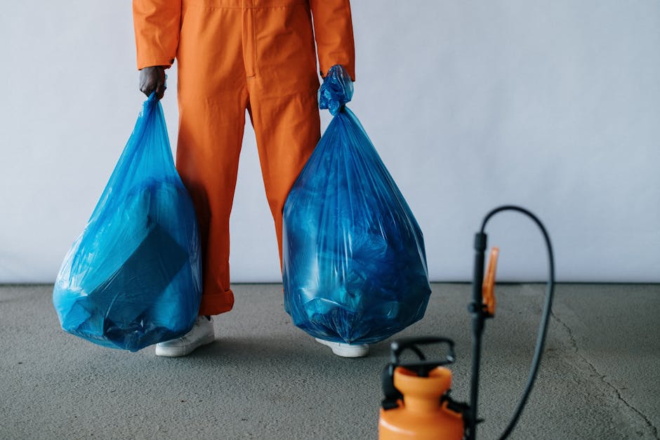 A person standing indoors on a grey carpeted floor, visible only from the waist down, wearing orange trousers and white sneakers, holding two large blue plastic rubbish bags filled with waste, one in each hand, with arms slightly bent. The bags are transparent in parts, showing crumpled recyclable materials inside. The background features a plain white wall, suggesting an indoor setting for waste collection or clearance. The scene exemplifies private rubbish disposal and on-site clearance activities, with the individual ready for waste removal services such as those offered by Rubbish Clearance Docklands, providing an informal but clear view of waste collection and disposal procedures fitting within the context of independent rubbish removal efforts.