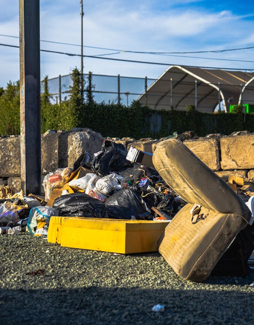 A mound of mixed waste materials on the ground beside a low stone wall, with black and transparent plastic garbage bags, a yellow plastic container, an old worn-up car tire, and assorted packaging and debris. The waste is situated outdoors on a gravel surface, with a utility pole and overhead wires visible in the background. Behind the stone wall, there is a hedge and a large metal structure, possibly a sports or industrial building, under a partly cloudy sky. The scene appears to convey an example of unmanaged rubbish accumulation, highlighting the importance of private rubbish clearance services such as those provided by Rubbish Clearance Docklands, especially in contexts of alternative waste handling or on-site clearance solutions.