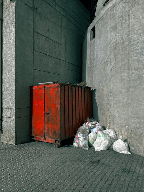 A large, industrial red metal skip with visible signs of rust and wear, positioned against a dark green and light grey building exterior on a paved street surface. The skip is rectangular with vertical ridges on its sides, and its top edge is slightly open, revealing a dark interior. Next to the skip, on the pavement, there are several white plastic bags filled with mixed rubbish, including clear bags that contain recyclable materials such as plastic bottles and other waste. The background features a modern, dark-colored wall with a small, rectangular window or vent near the top, and part of a light grey concrete wall to the right. The scene appears to be an urban alleyway or service access point, with natural daylight illuminating the area, typical of on-site rubbish collection or private waste disposal setups. This setup aligns with private rubbish clearance services, like those offered by Rubbish Clearance Docklands, for managing waste outside of council collection points, emphasizing the importance of proper disposal methods for bulky and mixed waste.